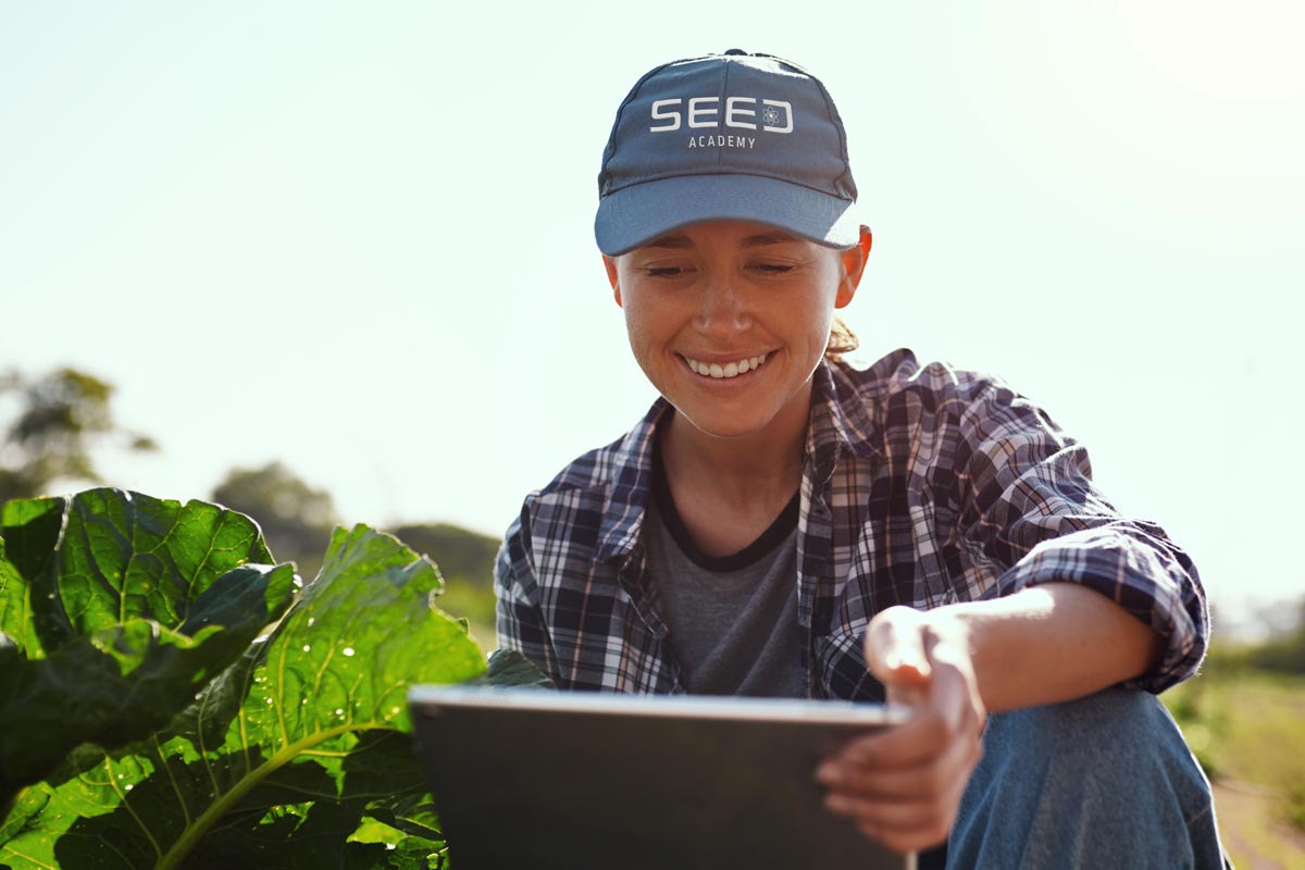 youn woman in an agricultural field looking at a digital tablet and wearing a SEED Academy logo cap